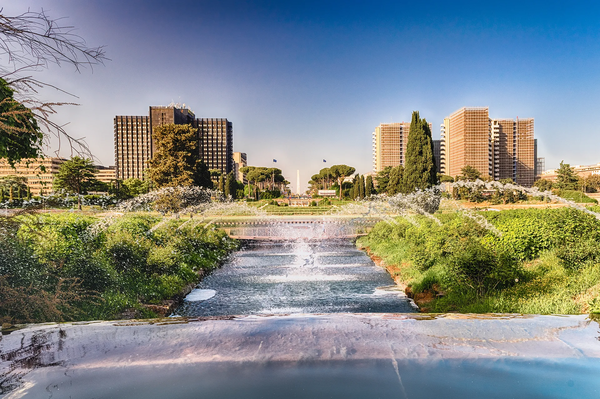 Roma, zona EUR: fontana con getti d'acqua in un ampio giardino urbano con edifici alti sullo sfondo sotto un cielo azzurro limpido.