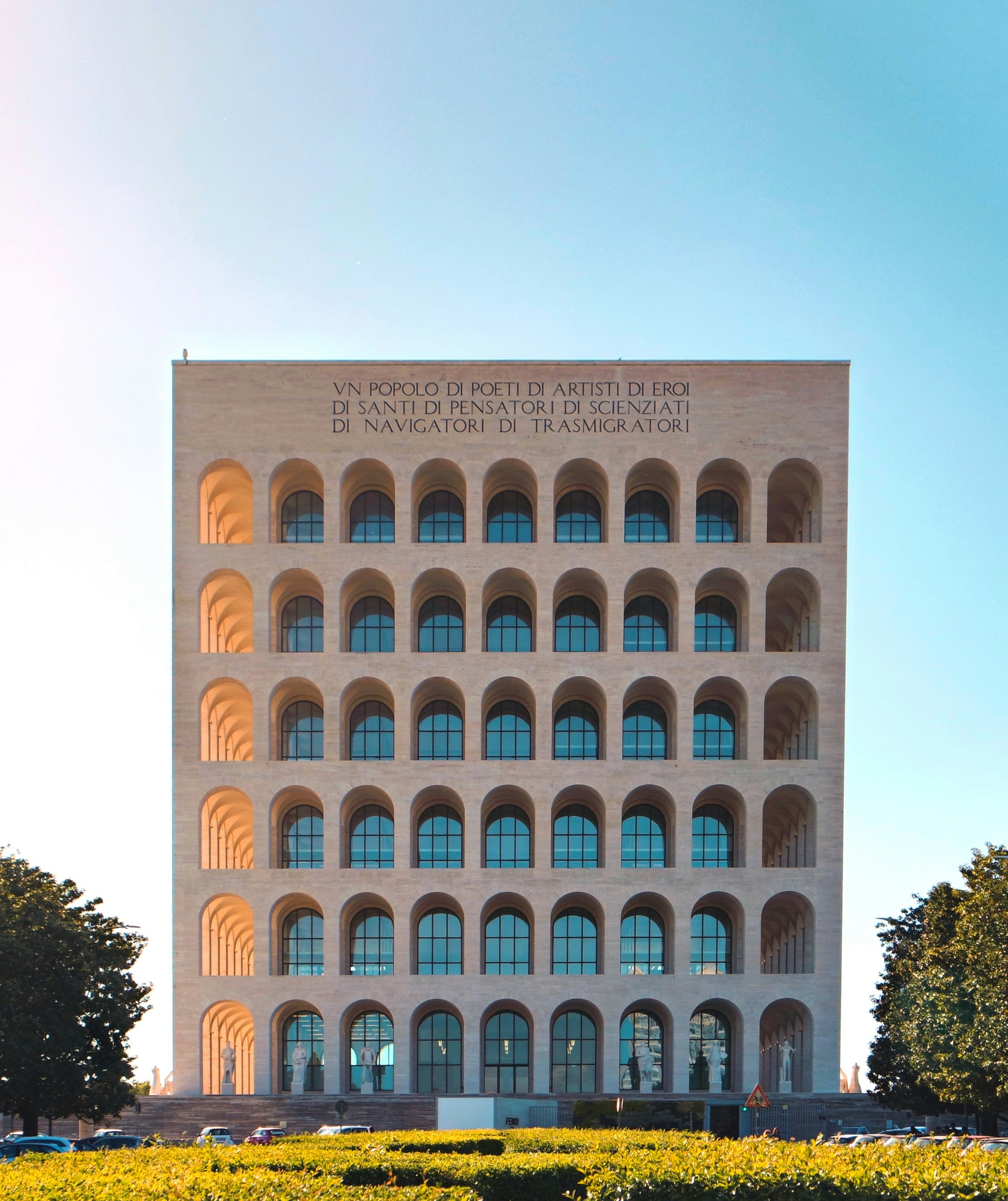 Palazzo della Civiltà Italiana a Roma con facciata a sei file di archi simmetrici e iscrizione in cima, circondato da vegetazione e parcheggio.